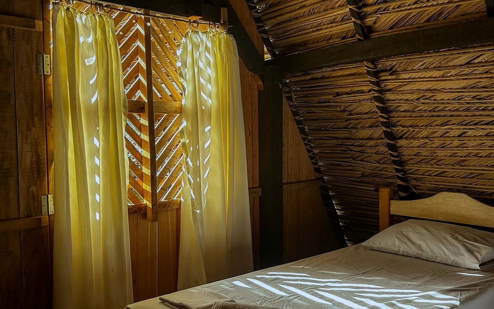Cama de solteiro junto à janela com cortina, luz entrando pelas venezianas
